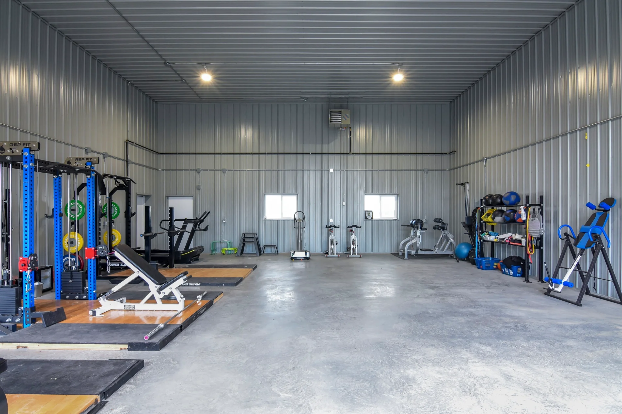 Gym equipment fills the interior of a post frame building and is finished with metal cladding and a concrete floor.