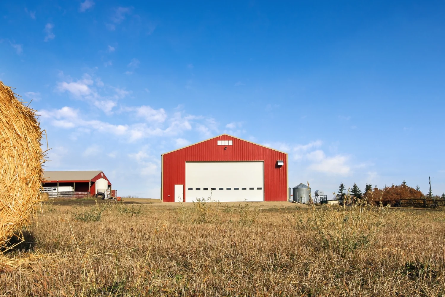 A hay bale in the foreground with a red farm shop building nearer to the background.