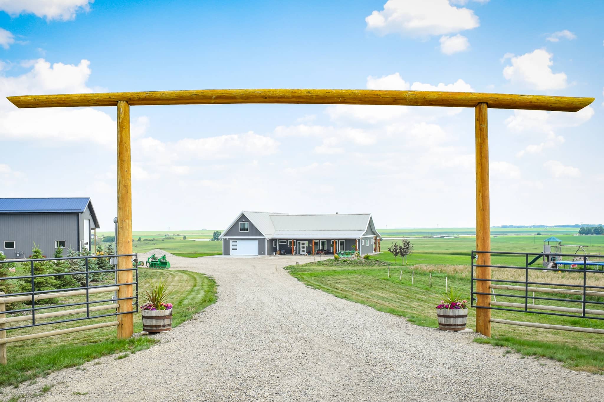 The arch at the end of a rural driveway frames a grey post frame bungalow.