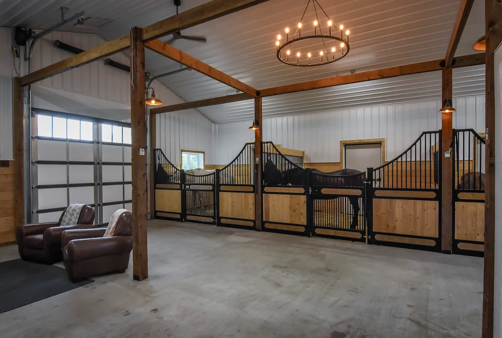 Carriage doors, windows, and lighting fixtures create a brightly lit pole barn interior for horses